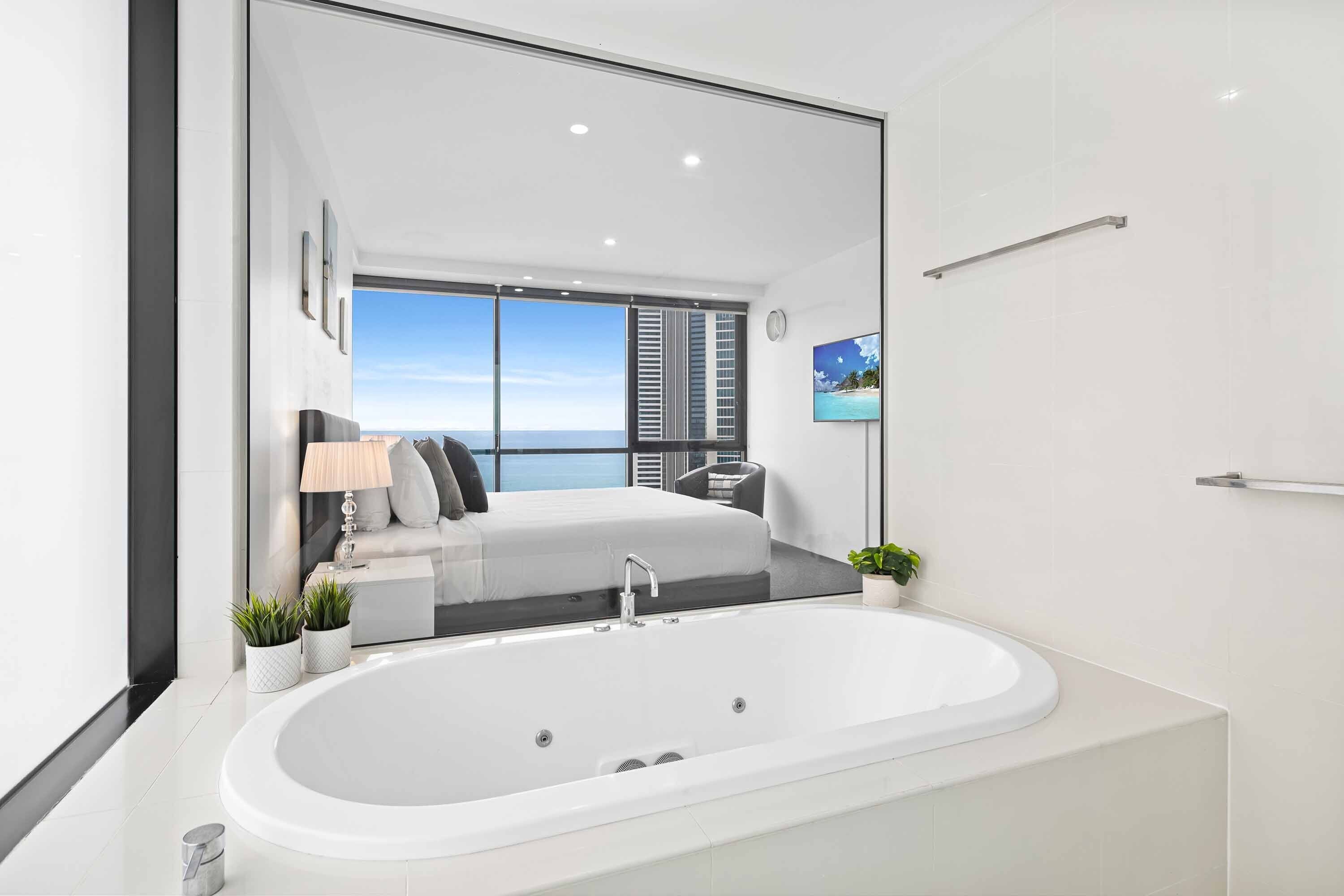 A modern bathroom with a large bathtub, a mirror, and a view of a balcony with a bed and plants.