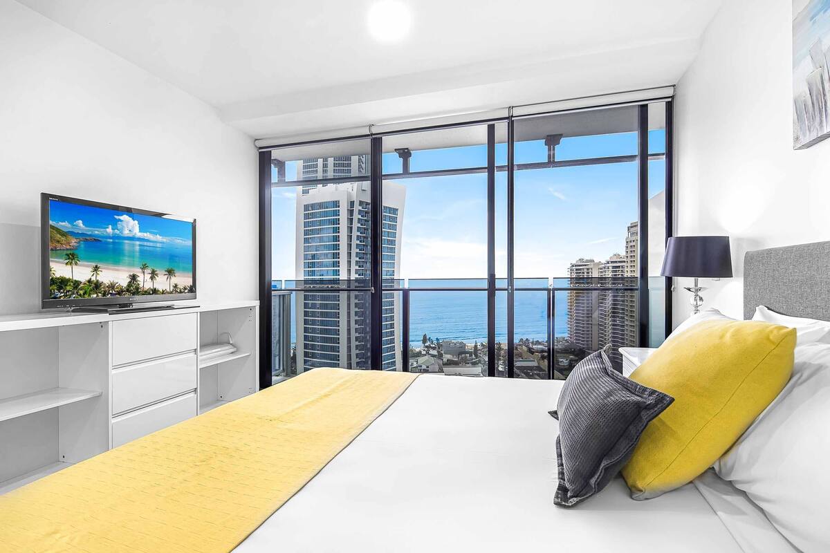 A modern hotel room with a large window, a bed with yellow and gray pillows, a TV on a white shelf, and a city view.