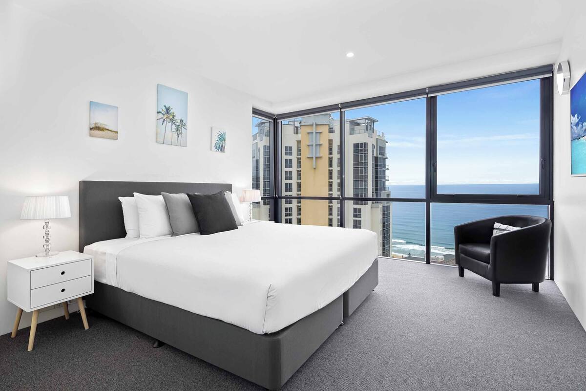 A modern bedroom with a large bed, a nightstand, a chair, and a view of the ocean through floor-to-ceiling windows.