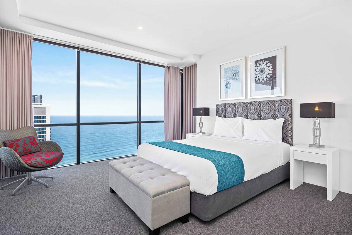 A modern hotel room with a large bed, a chair, a bench, a nightstand, and a view of the ocean.
