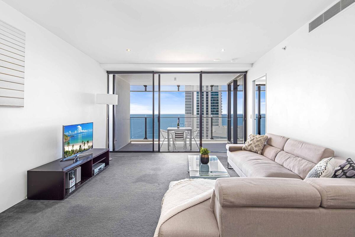 A modern living room with a large flat-screen TV, a beige sofa, a glass coffee table, and a view of the ocean through floor-to-ceiling windows.