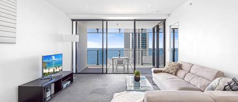 A modern living room with a large flat-screen TV, a beige sofa, a glass coffee table, and a view of the ocean through floor-to-ceiling windows.