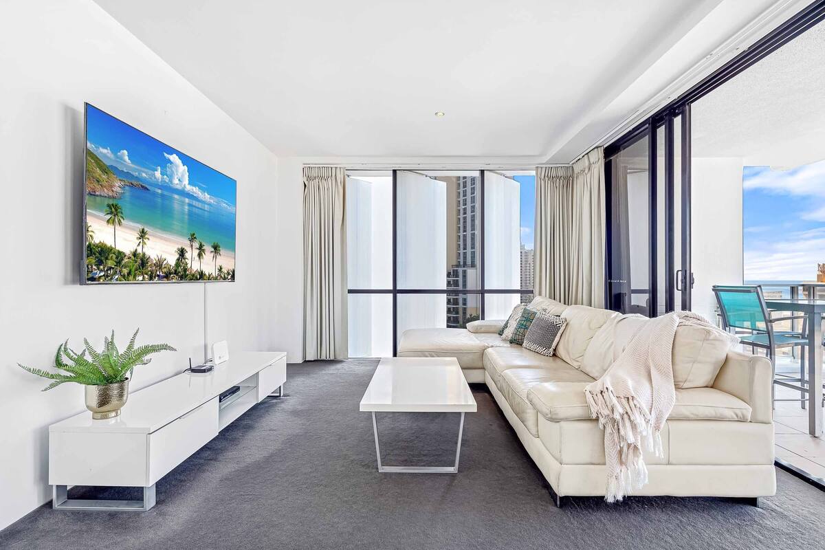 A modern living room with a large window, a sofa, a coffee table, and a TV displaying a beach scene.