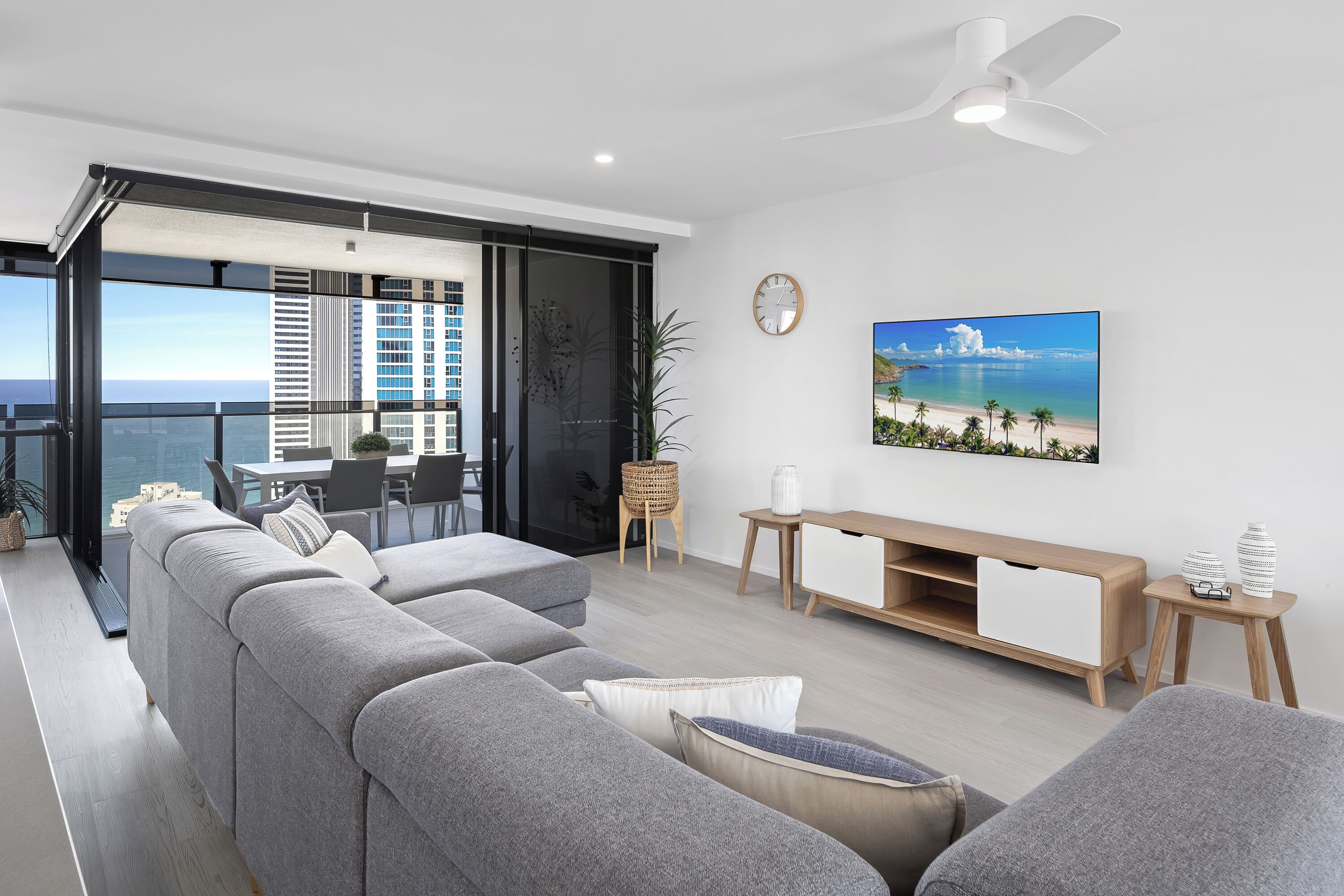 Een moderne woonkamer met een grijze hoekbank, een houten tv-meubel en een groot raam met uitzicht op een stadsbeeld.