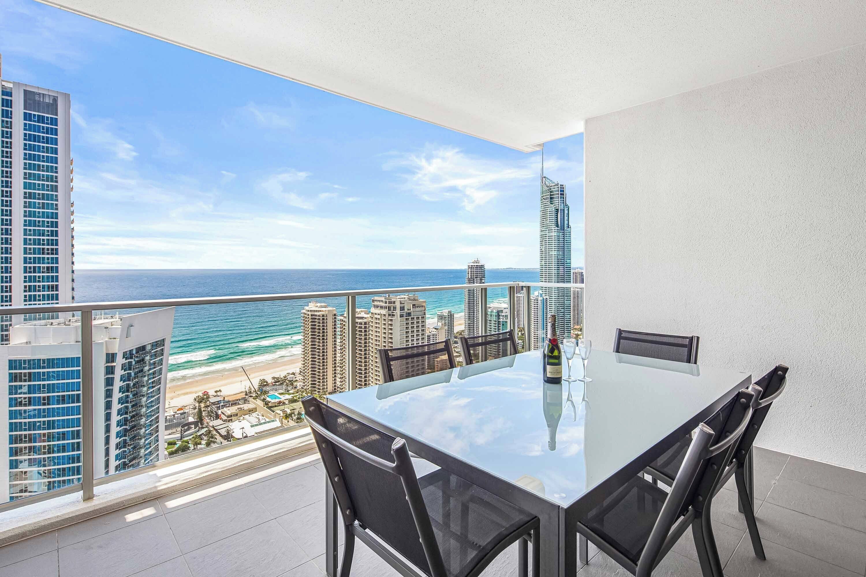A balcony with a glass table and chairs overlooking a beach and ocean.