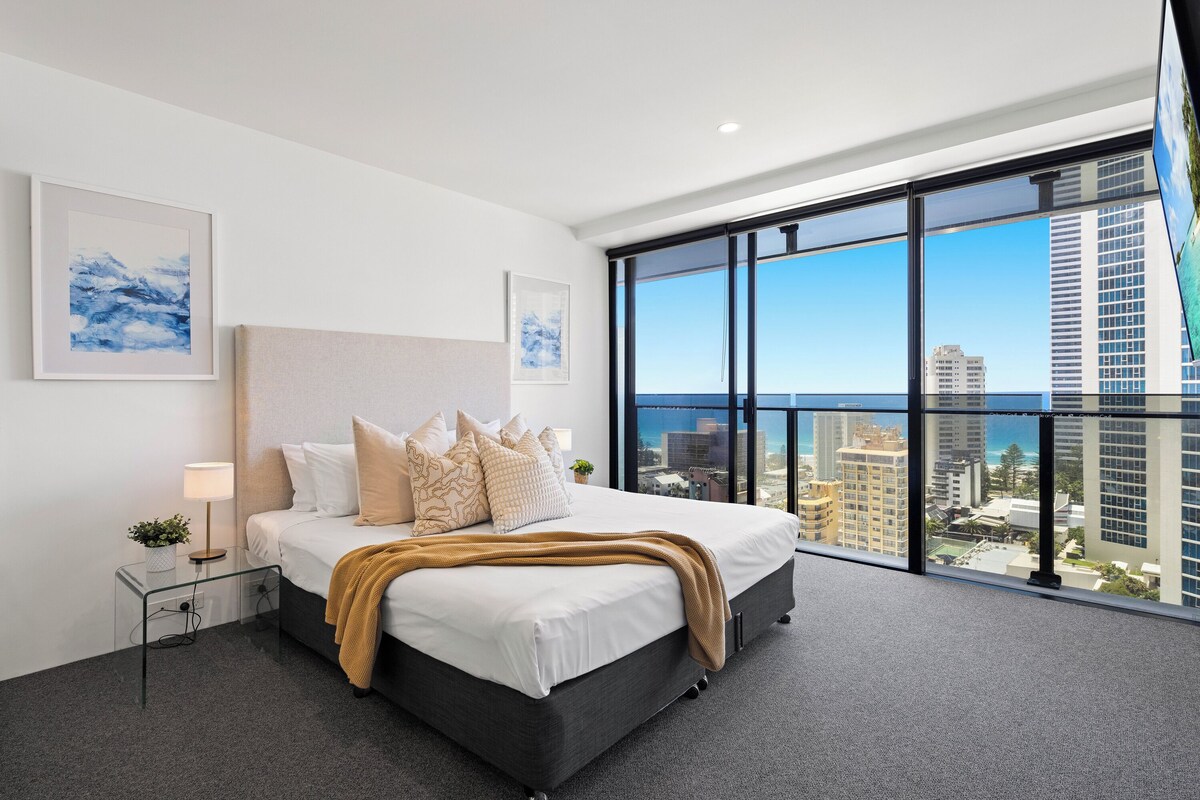 A modern hotel room with a large bed, a nightstand, a lamp, and a view of the city through floor-to-ceiling windows.