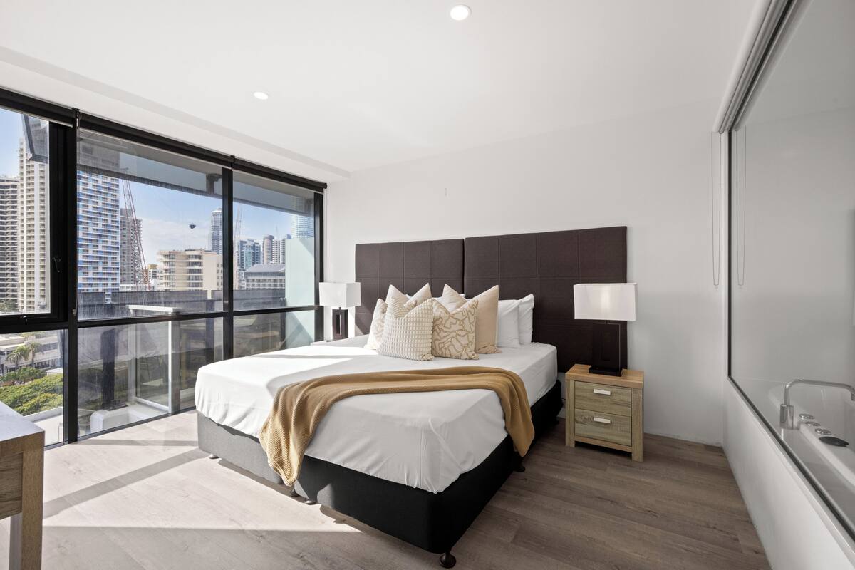 A modern bedroom with a large bed, bedside tables, a view of the city through floor-to-ceiling windows, and a cityscape visible outside.