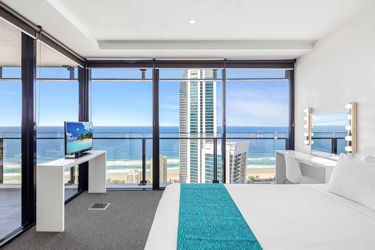 A modern hotel room with a large bed, a TV, a desk with a computer, and a bathroom with a mirror. The room has large windows offering a view of the ocean and tall buildings.