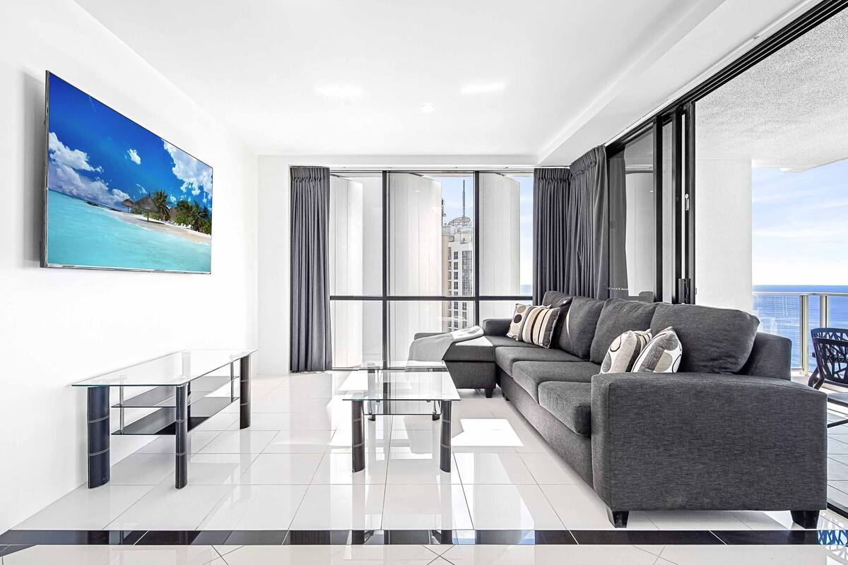 A modern living room with a large sofa, a glass coffee table, and a flat-screen TV mounted on the wall.