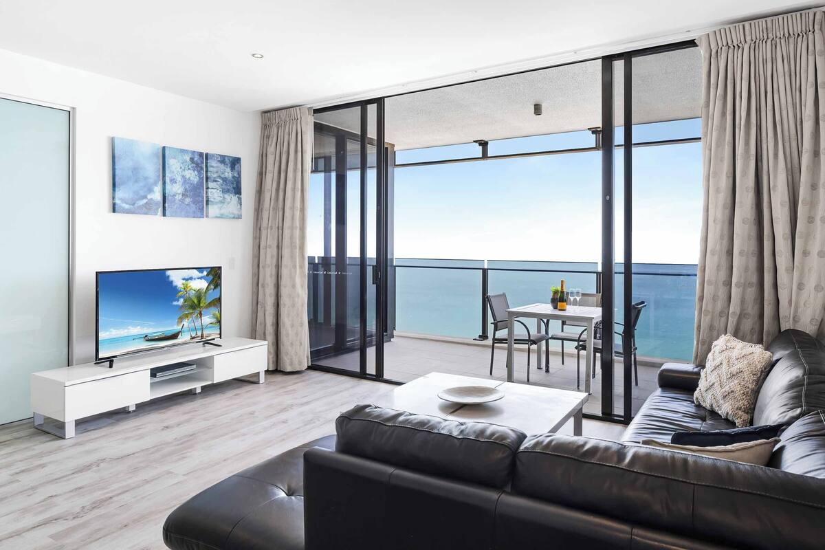 A modern living room with a large TV, a sofa, a coffee table, and a balcony with a view of the sea.