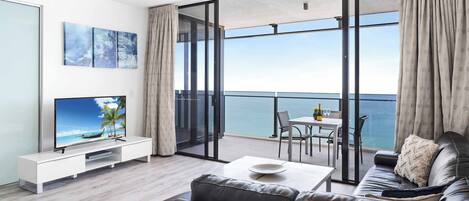 A modern living room with a large TV, a sofa, a coffee table, and a balcony with a view of the sea.