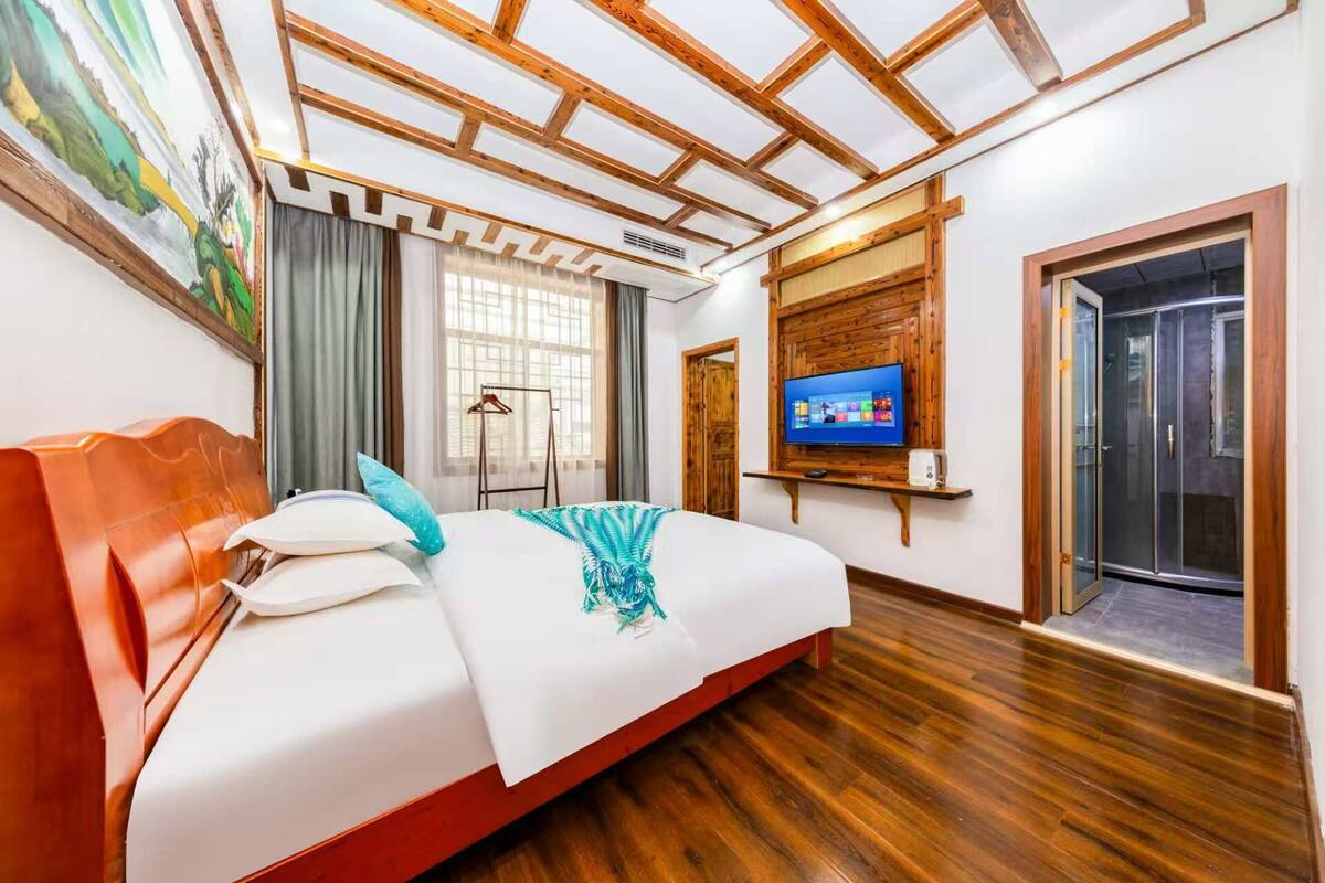 Une chambre d’hôtel avec un plafond en bois, un lit avec une literie blanche et un plaid bleu, un téléviseur fixé au mur et une porte donnant sur une salle de bain.