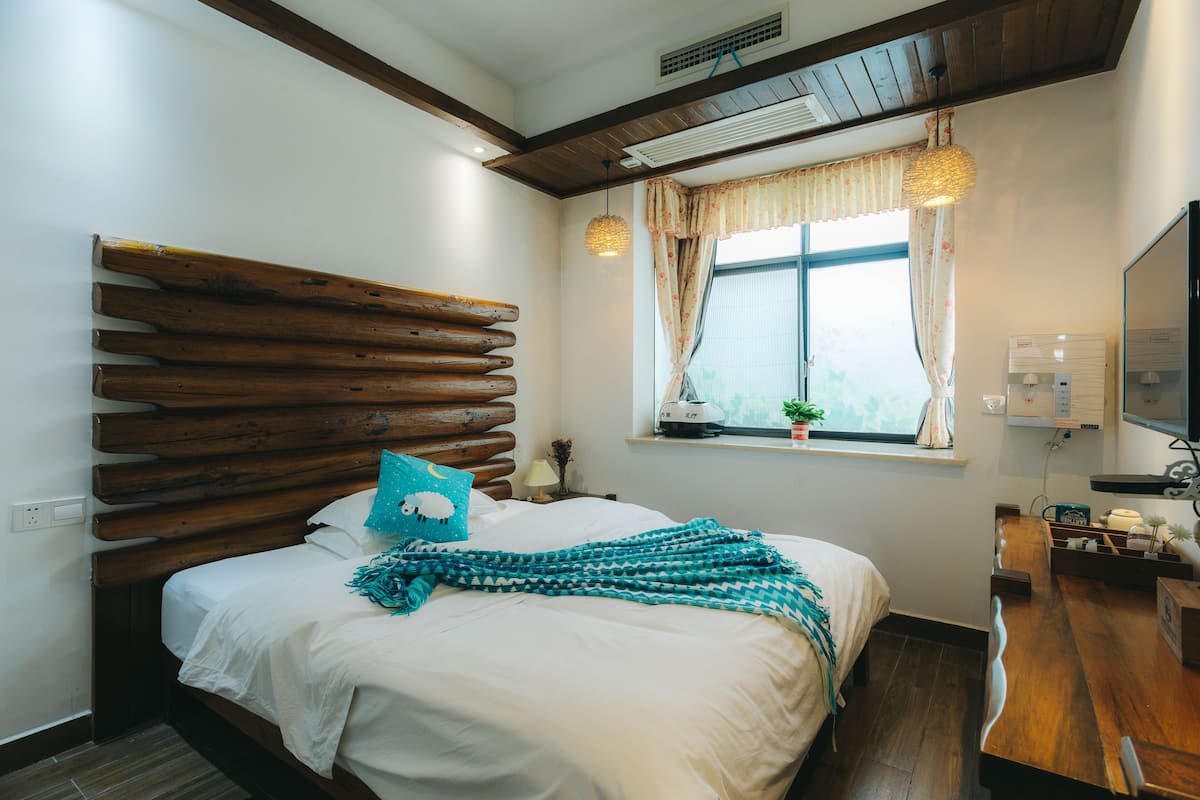 Une chambre avec une tête de lit en bois, un lit avec une literie blanche, un plaid turquoise, une fenêtre avec des rideaux, une table de chevet avec une lampe et un téléviseur fixé au mur.