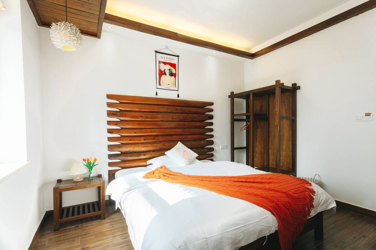Une chambre à coucher avec une tête de lit en bois, un lit avec une literie blanche et un plaid orange, une petite table de chevet avec une lampe et des fleurs, et une affiche accrochée au mur.