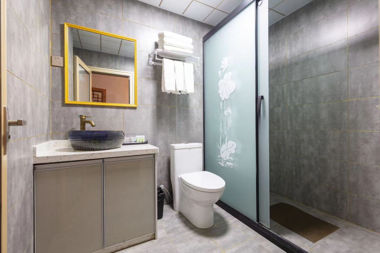 A modern bathroom with a grey tiled wall, a white toilet, a glass shower enclosure, and a sink with a decorative bowl.