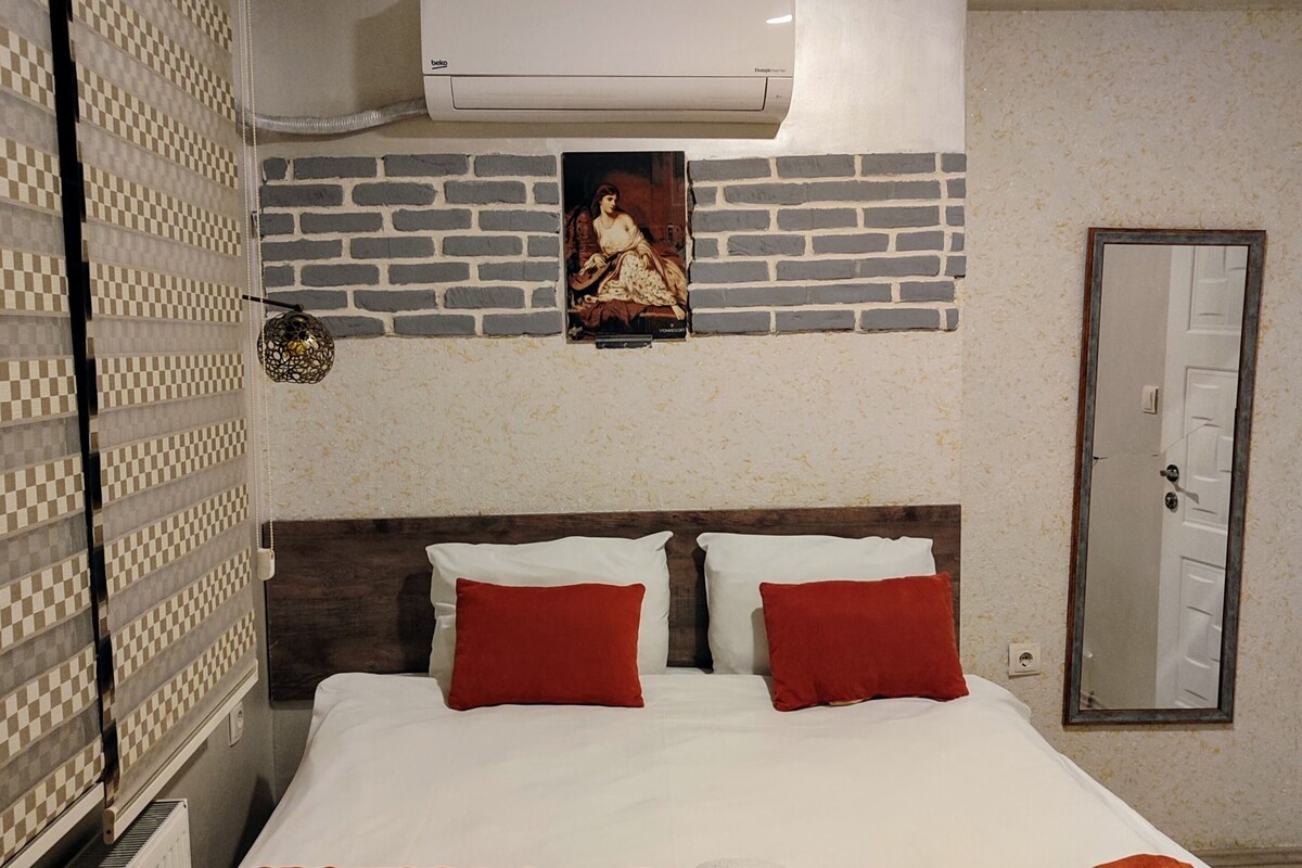 A hotel room with a bed, red pillows, a white blanket, and a folded towel on the bed.