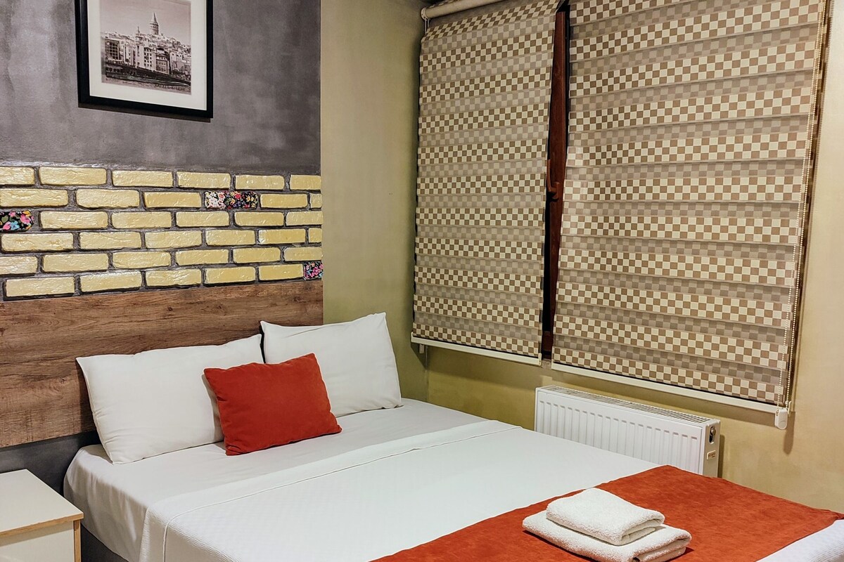 A neatly made bed with white and red bedding, a wooden headboard, and a checkered blind covering a window.