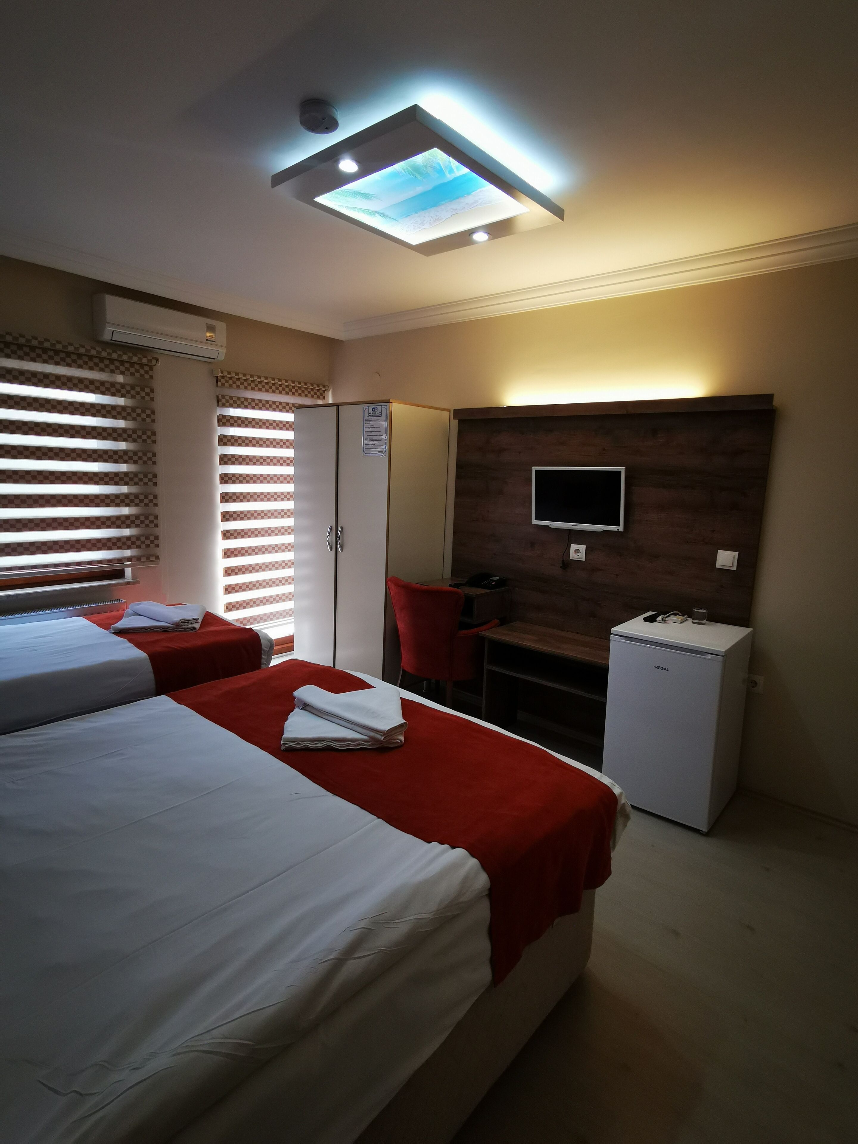 A hotel room with two beds, a TV mounted on a wooden panel, a refrigerator, and a ceiling light fixture.