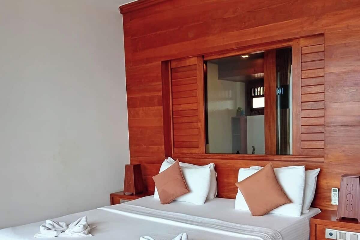 Quarto de hotel com cabeceira de madeira, uma cama grande com lençóis brancos e uma mesa de cabeceira com relógio.
