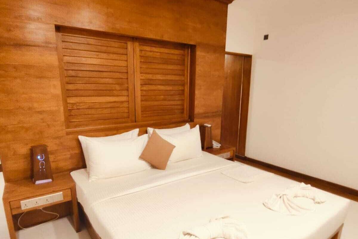 Quarto de hotel com cabeceira de madeira, cama com roupa de cama branca, mesa de cabeceira e criado-mudo de madeira.