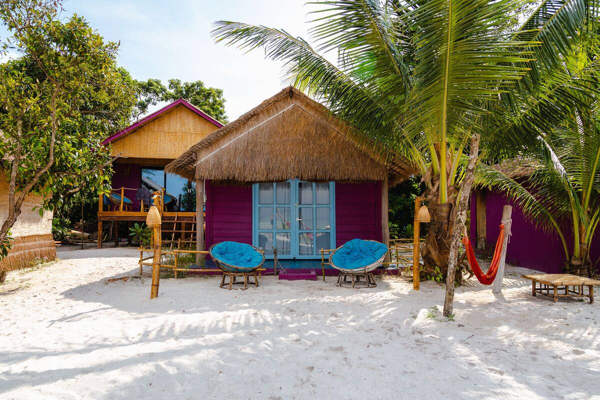 A beach house with a thatched roof, two blue cushioned chairs, and a hammock.