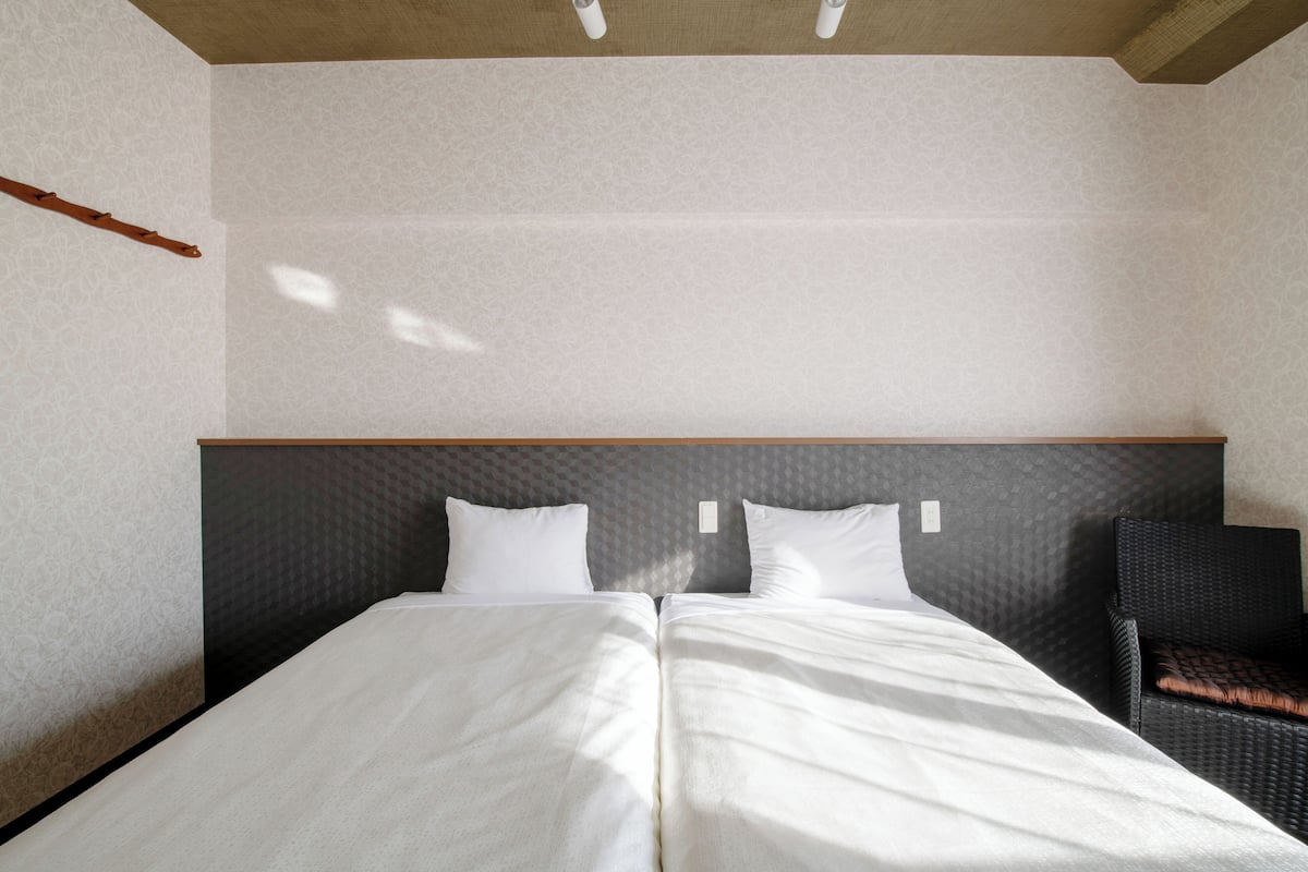Un lit double avec du linge de lit blanc, dans une chambre dotée d’une tête de lit grise à motifs et d’une chaise en osier.