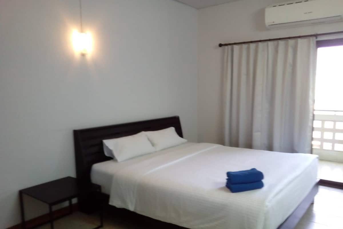 Habitación de hotel con una cama individual, una mesita de noche pequeña, aire acondicionado en la pared y una ventana con cortinas.