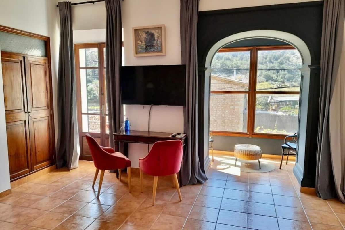 A room with a wooden desk, two red chairs, a television, and a view of a balcony with a stone wall and greenery.