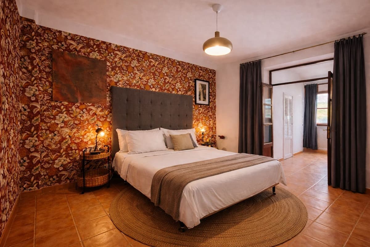 A bedroom with a bed, a headboard, a wall-mounted picture, a bedside lamp, and a view of the outside.