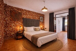 Un dormitorio con cama, cabecera, cuadro en la pared, lámpara de noche y vista al exterior.