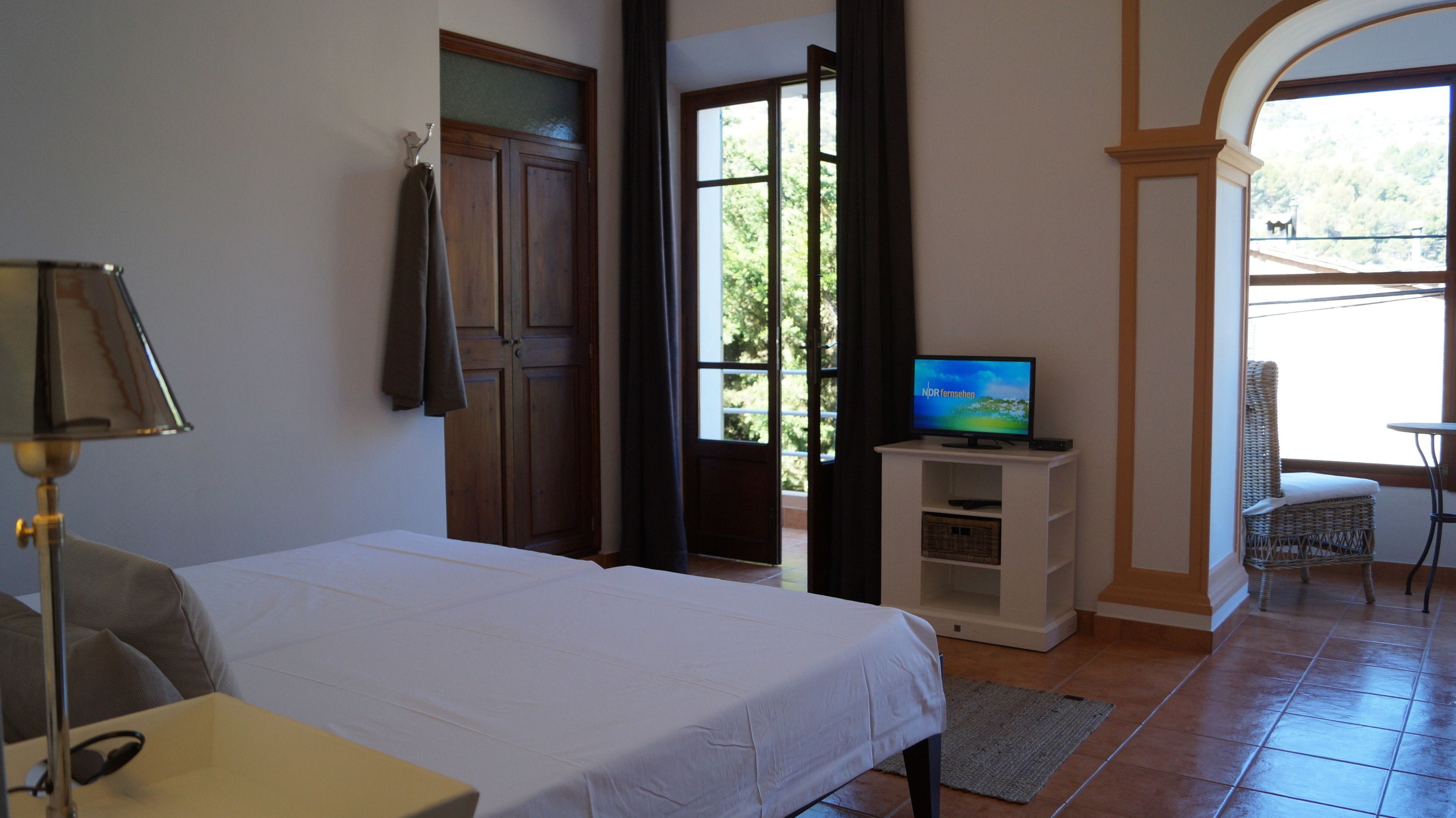 A bedroom with a bed, a television on a stand, a lamp, a door, and a view of the outdoors.