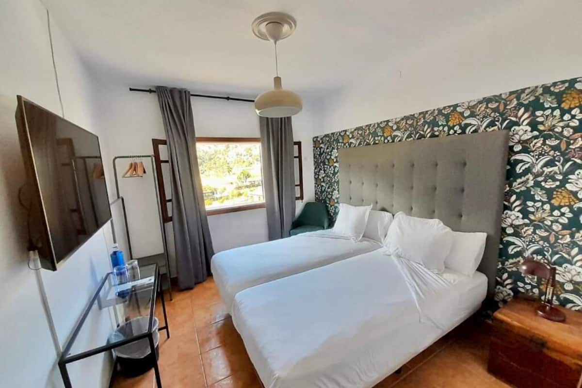 A hotel room with two beds, a TV, a small table with a water bottle, and a window with curtains.