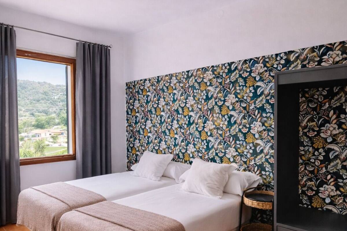 A double bed with a woven rug, a floral-patterned wall, and a view of greenery outside.