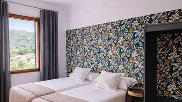 A double bed with a woven rug, a floral-patterned wall, and a view of greenery outside.