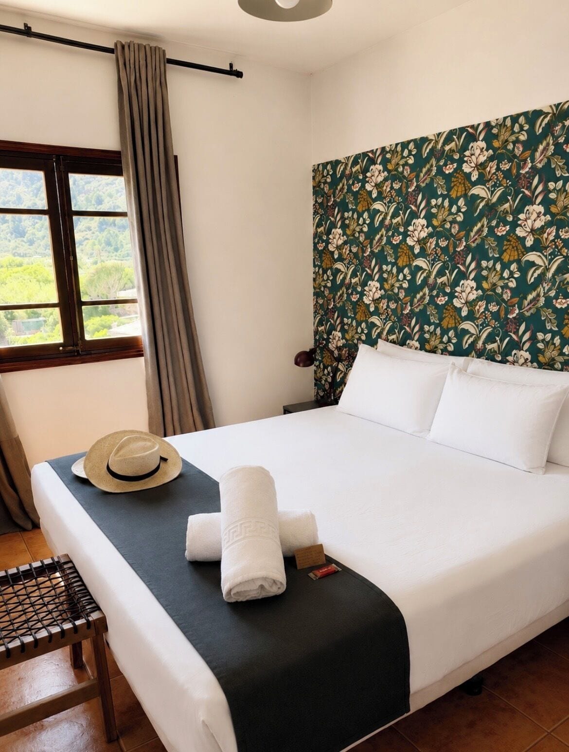 Una cama bien hecha con sábanas blancas, un sombrero y toallas sobre un cabecero estampado con motivos florales.