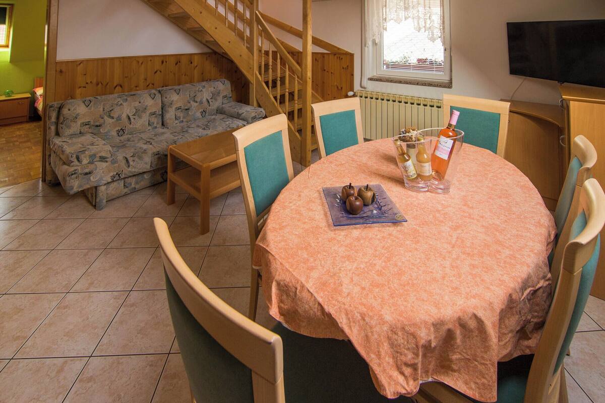 A dining area with a round table covered in a peach-colored tablecloth, surrounded by four chairs. A wooden staircase is visible in the background, leading to an upper level.