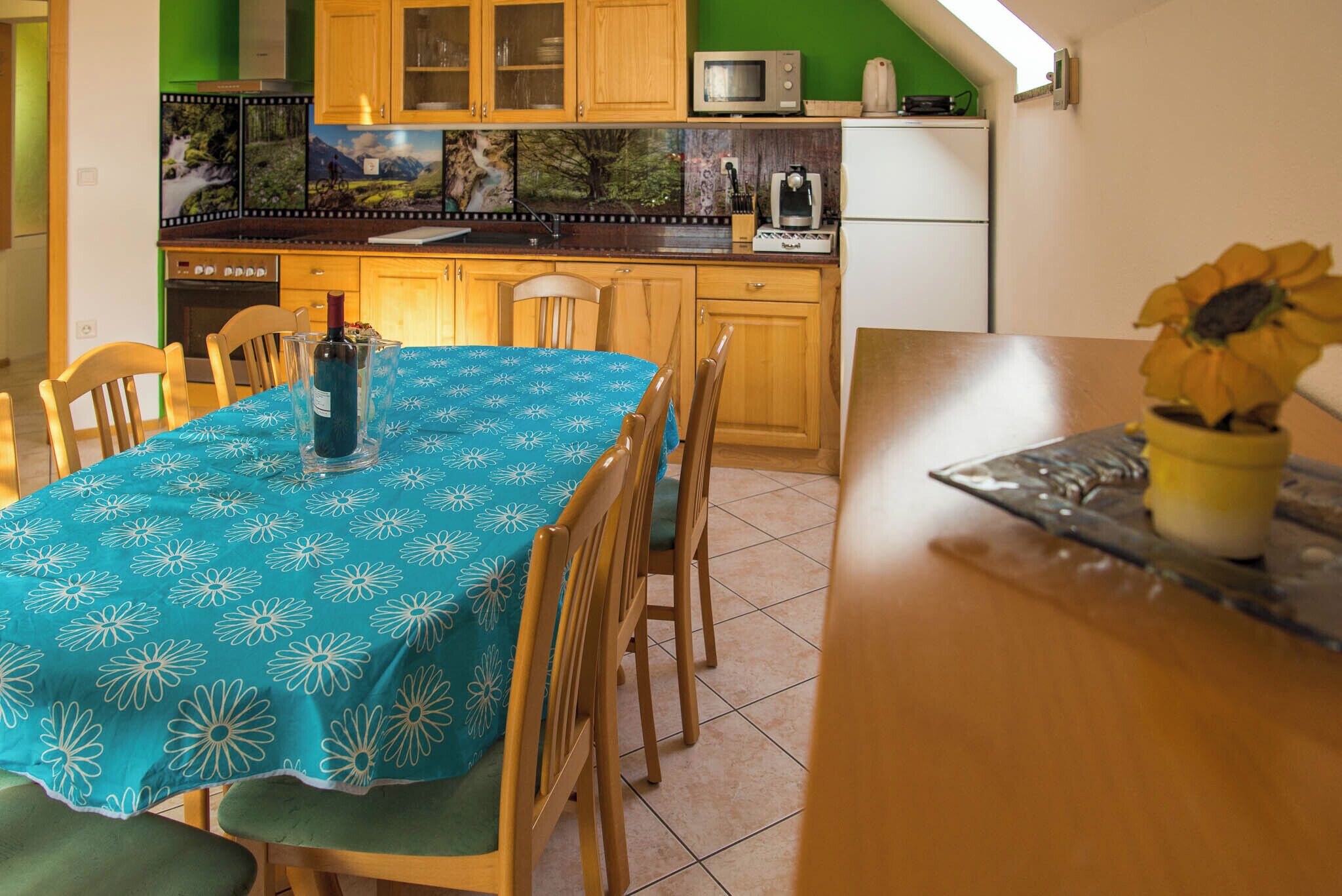A kitchen with wooden cabinets, a microwave, and a refrigerator. There is a dining table with a blue tablecloth and chairs. A vase with a sunflower is on the table.