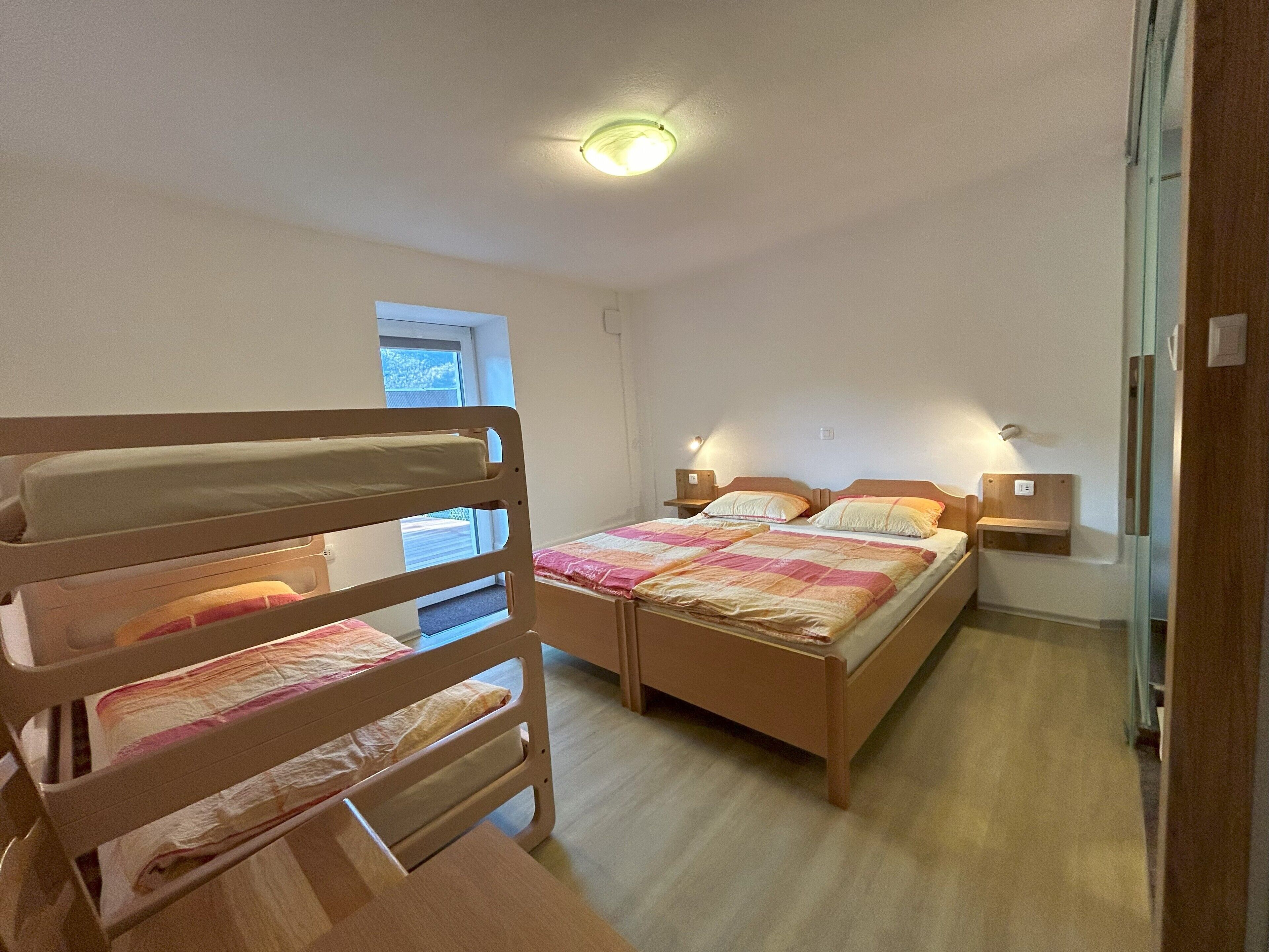 Ein Schlafzimmer mit zwei Einzelbetten, einem Etagenbett aus Holz, einem Nachttisch mit Lampe und einem Fenster mit Blick ins Freie.