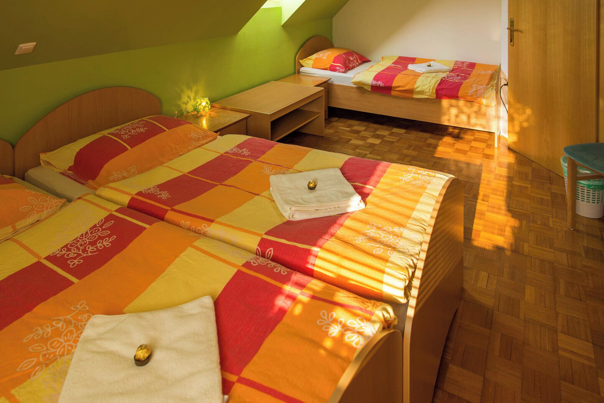 A room with two beds, each with a checkered bedspread and pillows. There is a wooden desk and a small table with a lamp. The room has a wooden floor and a green wall.