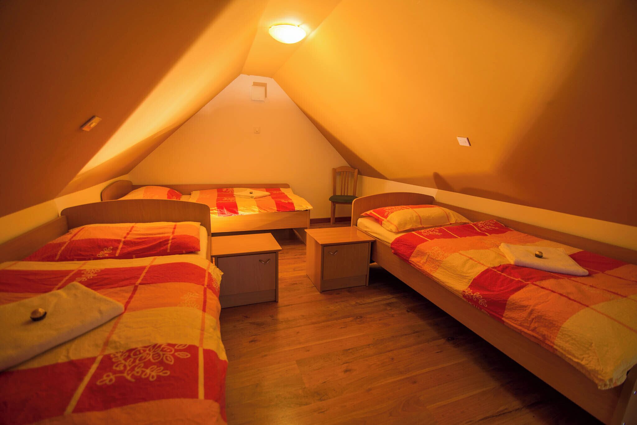 A small, triangular attic bedroom with two beds, each with a red and white quilt. There are bedside tables and a lamp.