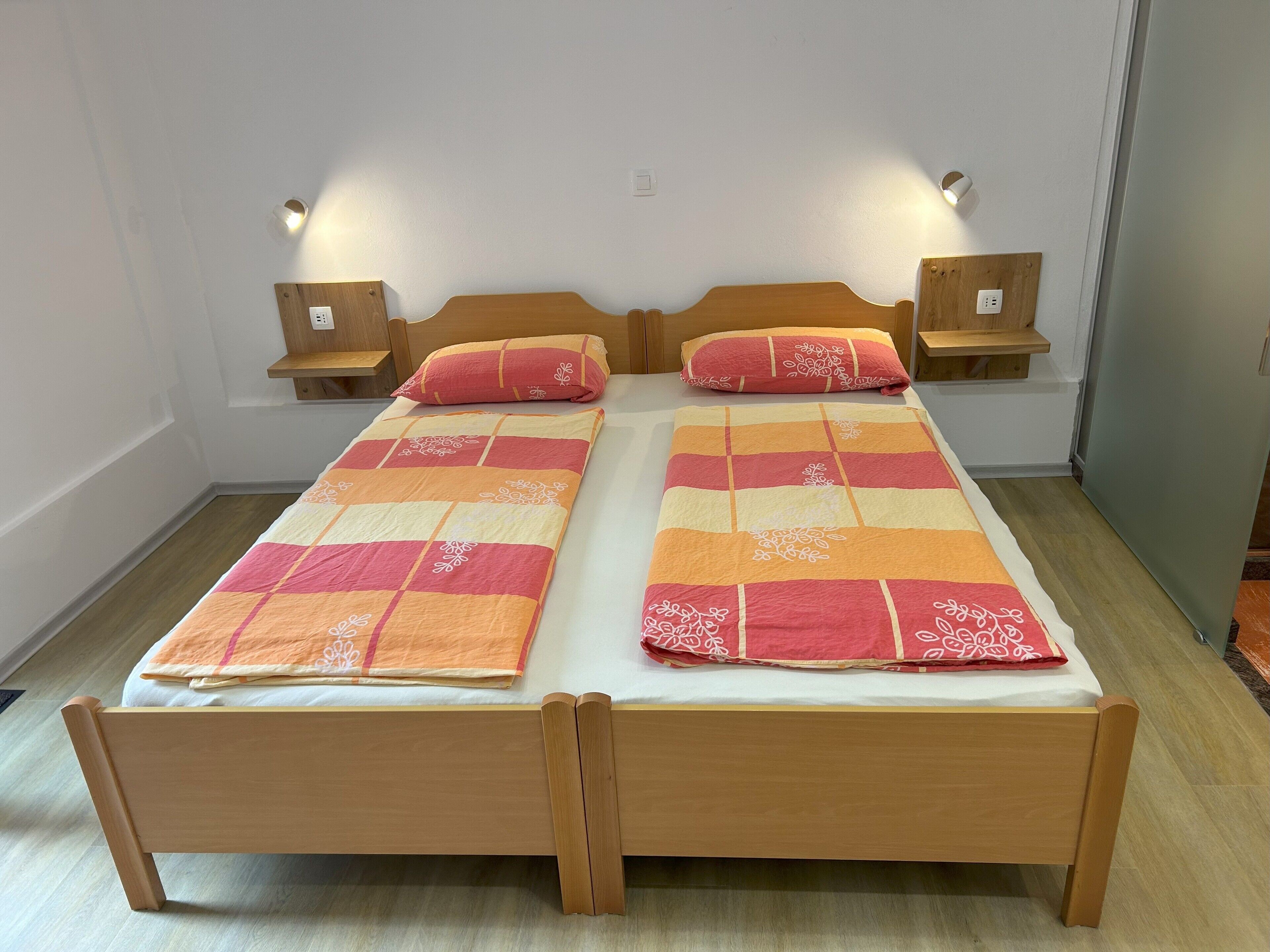 Una cama doble con estructura de madera, dos mesitas de noche con lámparas y un edredón de cuadros rojos y naranjas.