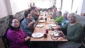 Un grupo de personas sentadas alrededor de una mesa de madera larga en una habitación con paredes de baldosas y una ventana.