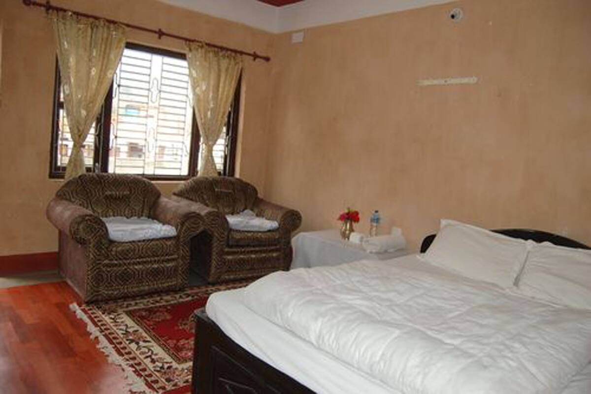 A bedroom with a large bed, two upholstered chairs, a small table with a vase, and a window with curtains.