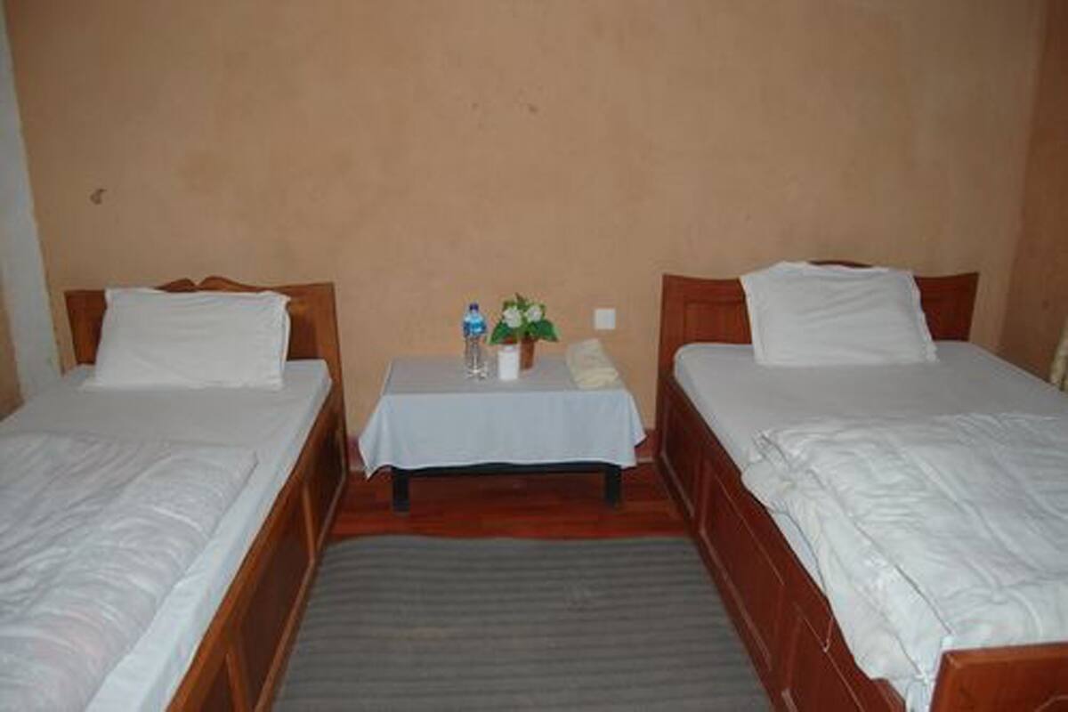 A room with two single beds, a small table with a tablecloth, a water bottle, and a potted plant.