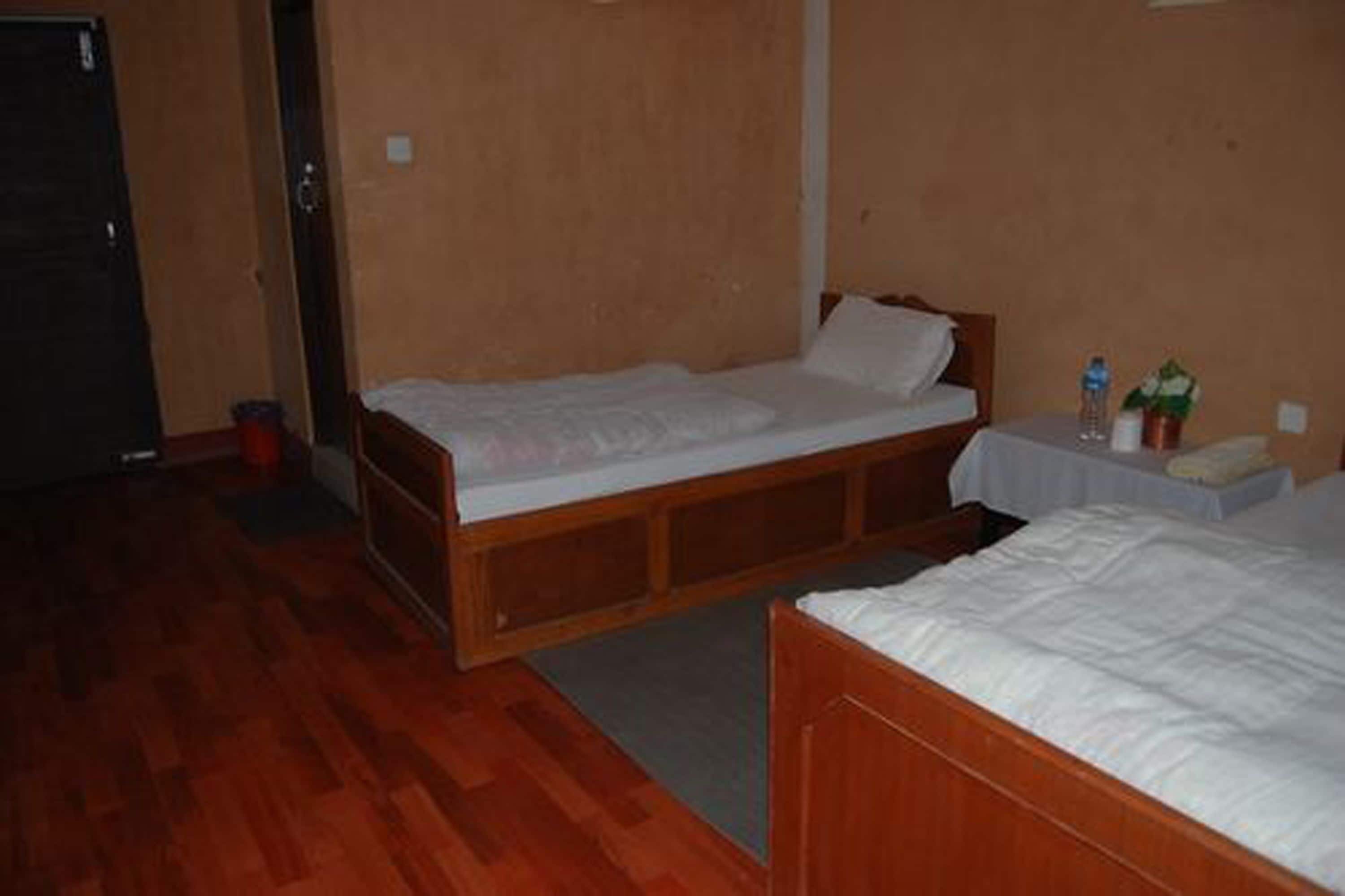 Une petite chambre d’hôtel, composée d’une seule pièce, avec un lit en bois, une petite table sur laquelle se trouvent une bouteille d’eau et une plante, et une porte.