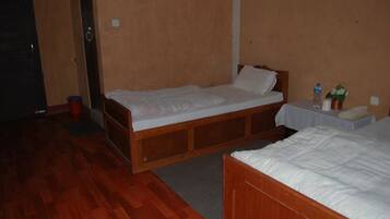 Une petite chambre d’hôtel, composée d’une seule pièce, avec un lit en bois, une petite table sur laquelle se trouvent une bouteille d’eau et une plante, et une porte.