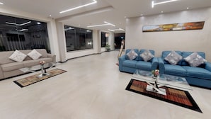Un saló modern amb un sofà beix i un sofà blau, amb coixins florals a tots dos. Hi ha taules de centre de vidre amb gerros i una planta en test. L'habitació té grans finestres i un quadre a la paret.
