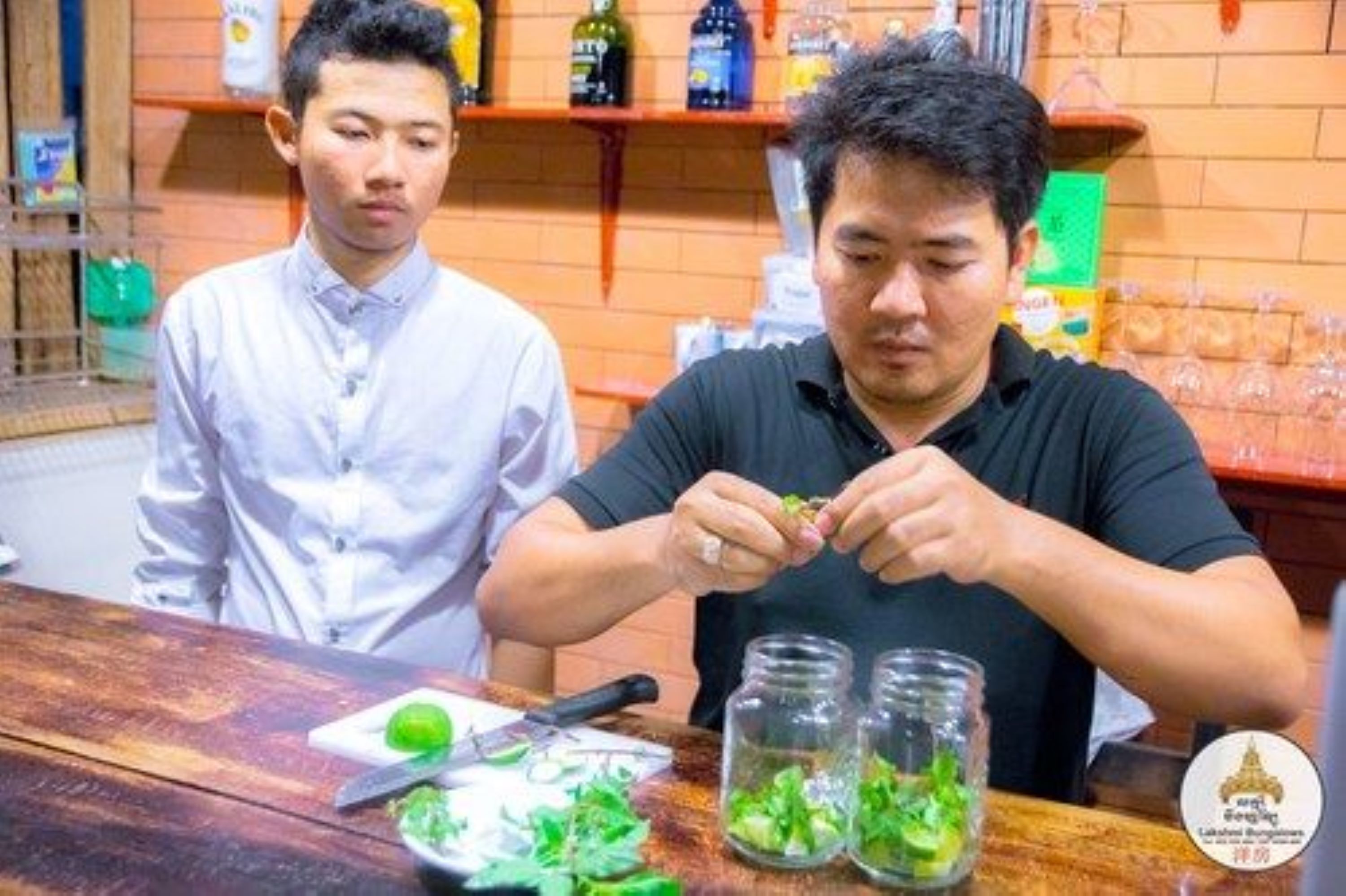 Two individuals preparing food at a counter with jars of green sauce and a cutting board with green leaves.