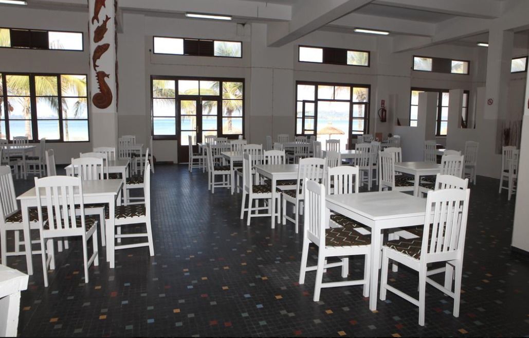 A spacious dining area with white tables and chairs, large windows with views of palm trees, and a tiled floor.