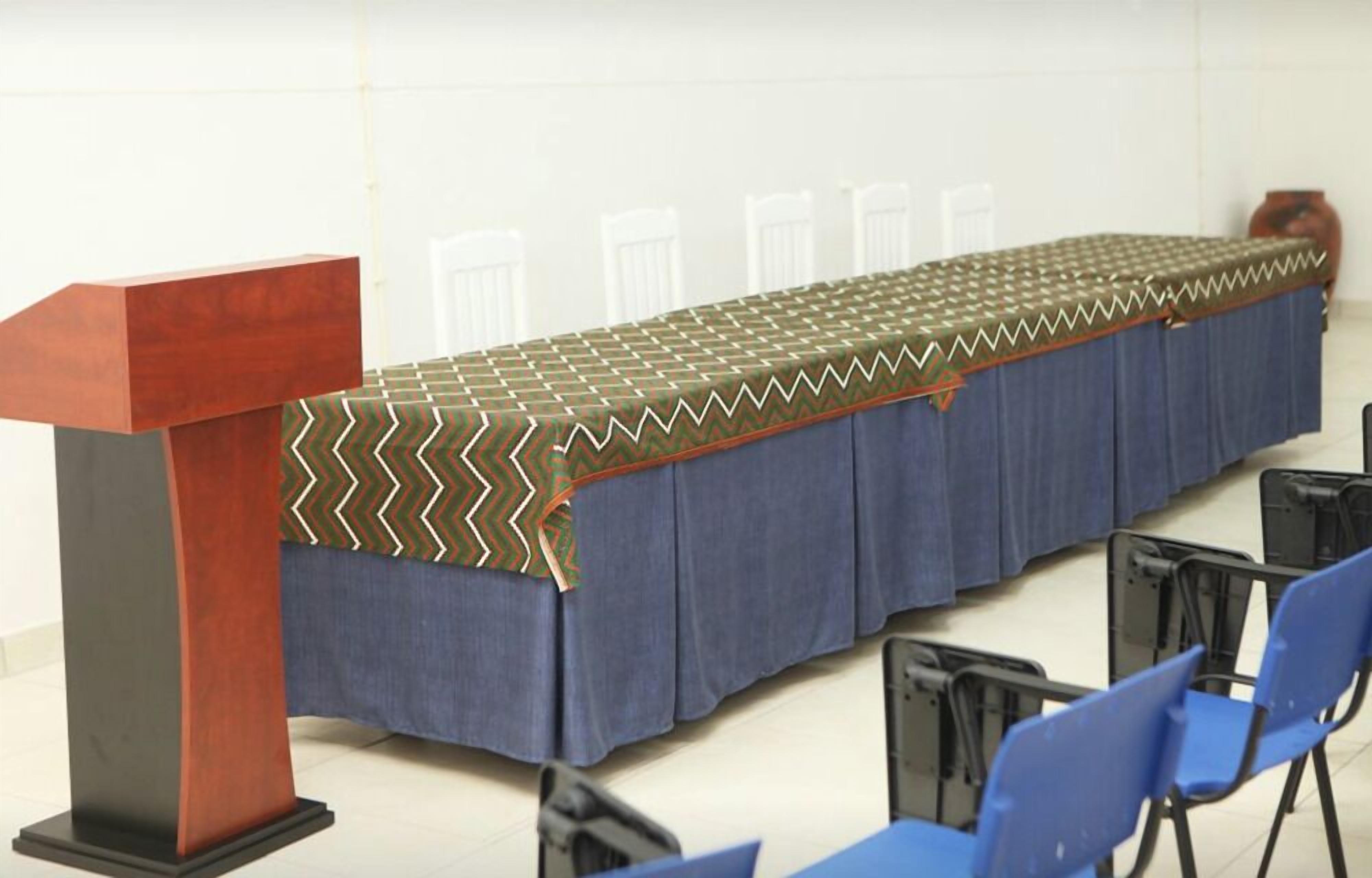 A conference room with a long table covered by a patterned tablecloth, blue chairs, and a wooden podium.